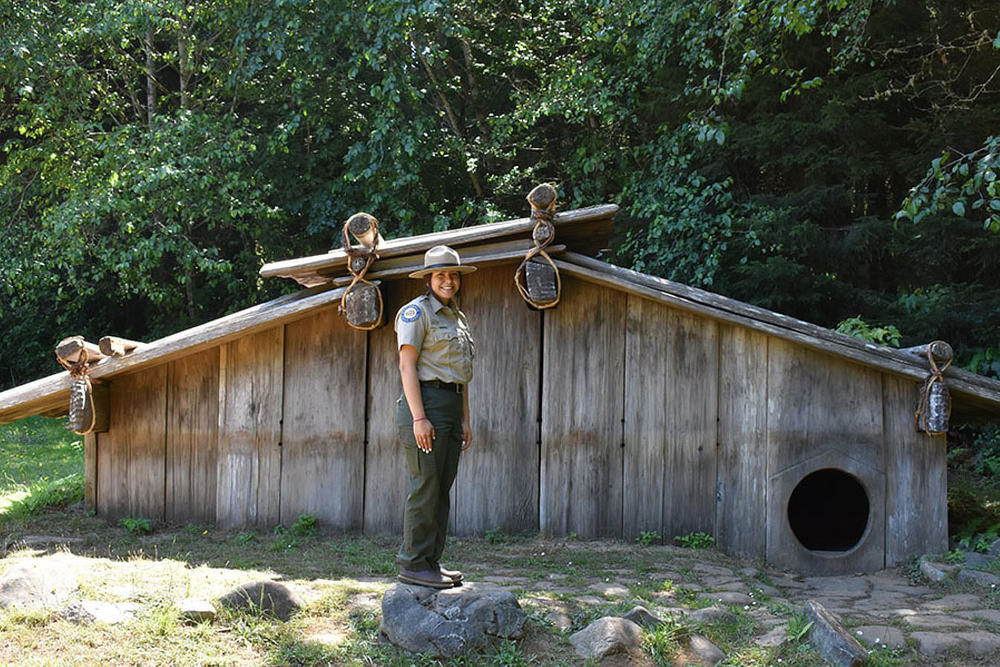 A female park ranger stands in front of a wooden hut and smiles at the viewer